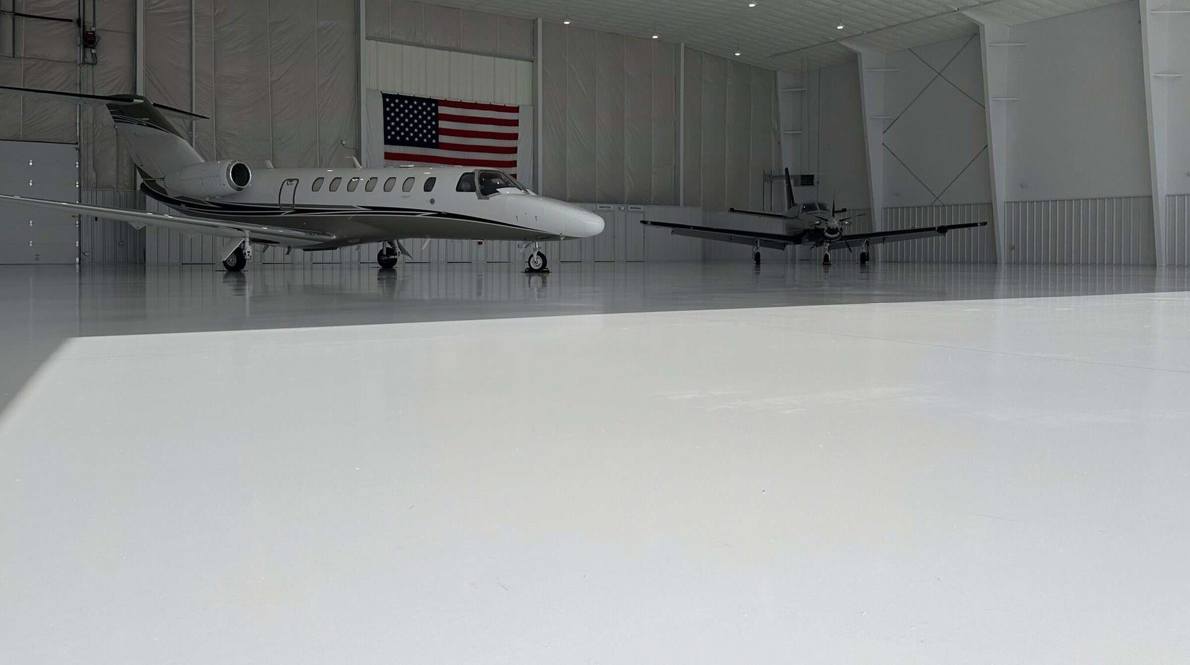 A Citation jet and a turboprop parked on the white epoxy floor inside the Shelton Aviation hangar, with the American flag on the back wall